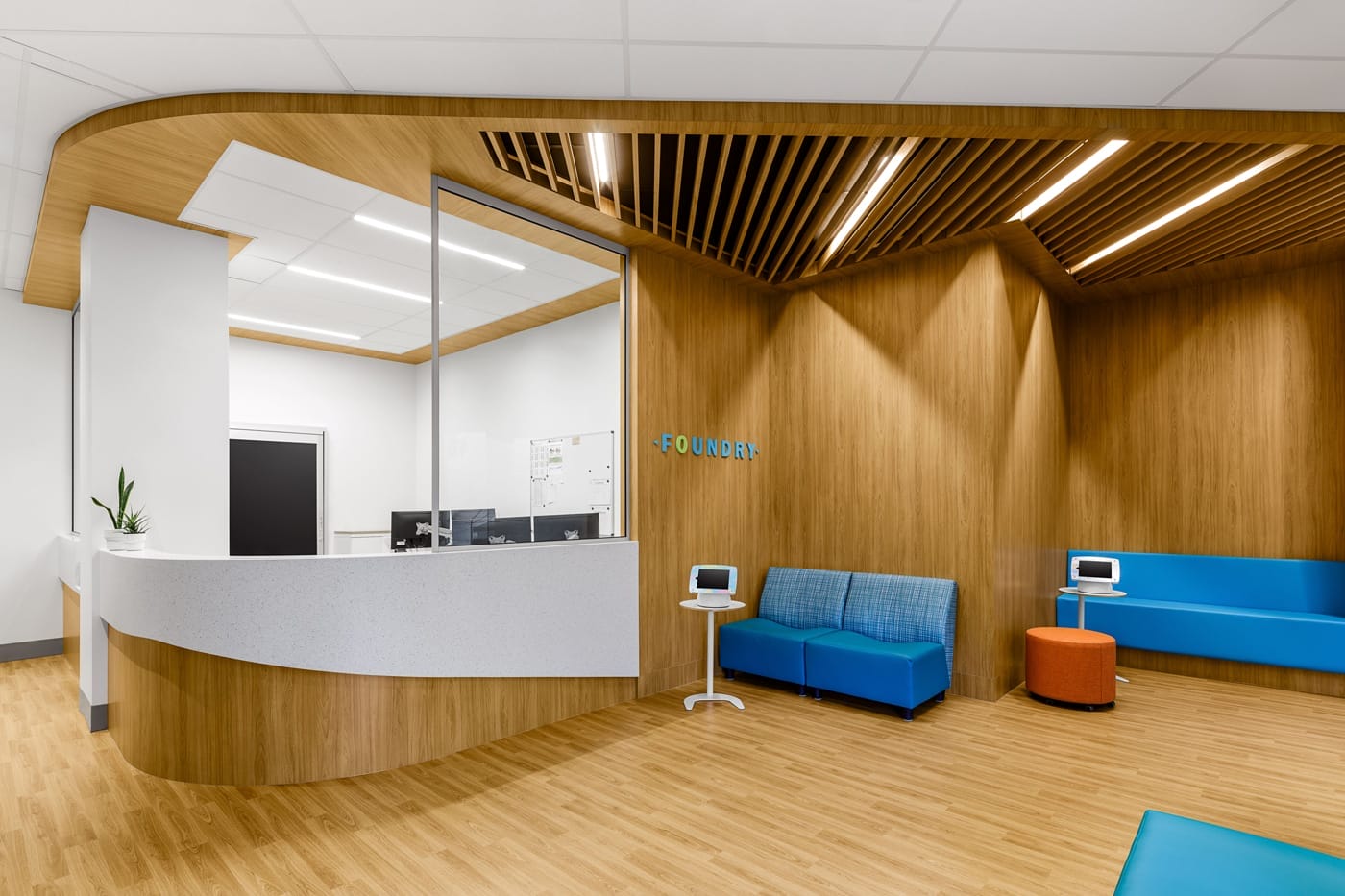 Bright, modern reception area at Foundry Richmond with a curved white reception desk, warm wood-paneled walls and ceiling slats, and vibrant blue seating, designed as a welcoming space for youth healthcare services.
